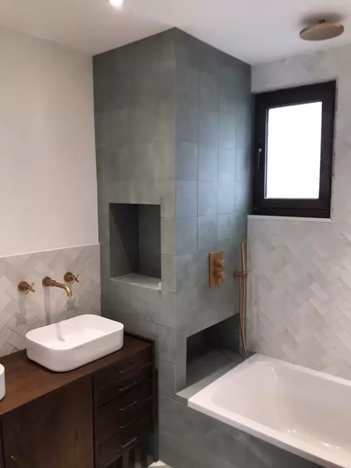 Salle de bain au carrelage vert avec niche, vasque posée sur meuble bois, robinetterie murale cuivrée et baignoire