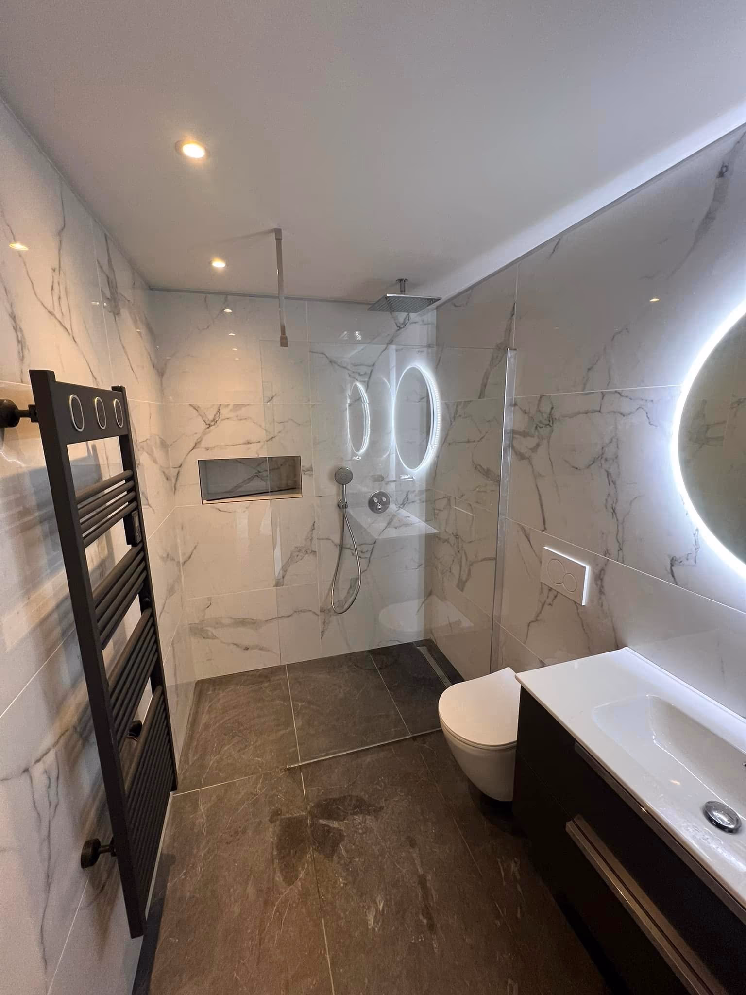 Salle de bain en marbre blanc avec miroirs ronds rétroéclairés, sèche-serviettes noir et douche vitrée
